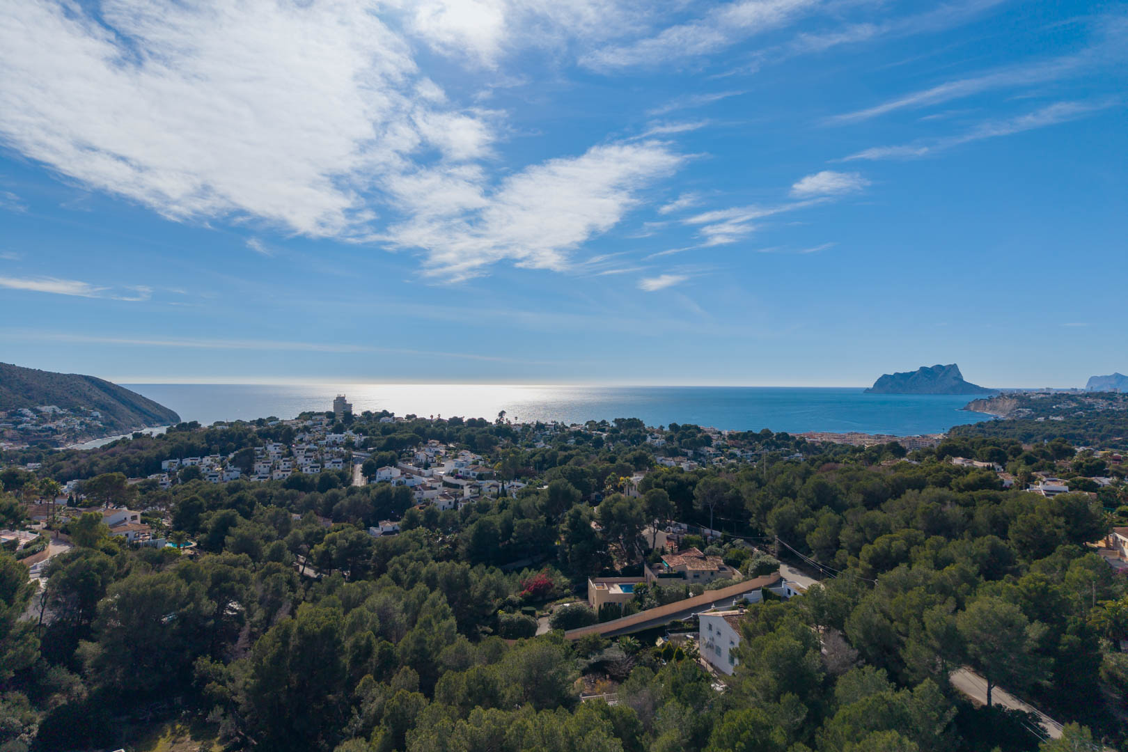 Panorama-Luftaufnahme der Küste von Moraira mit dichtem Pinienbestand, Wohnvillen und dem markanten Peñón de Ifach am Horizont des Mittelmeers.