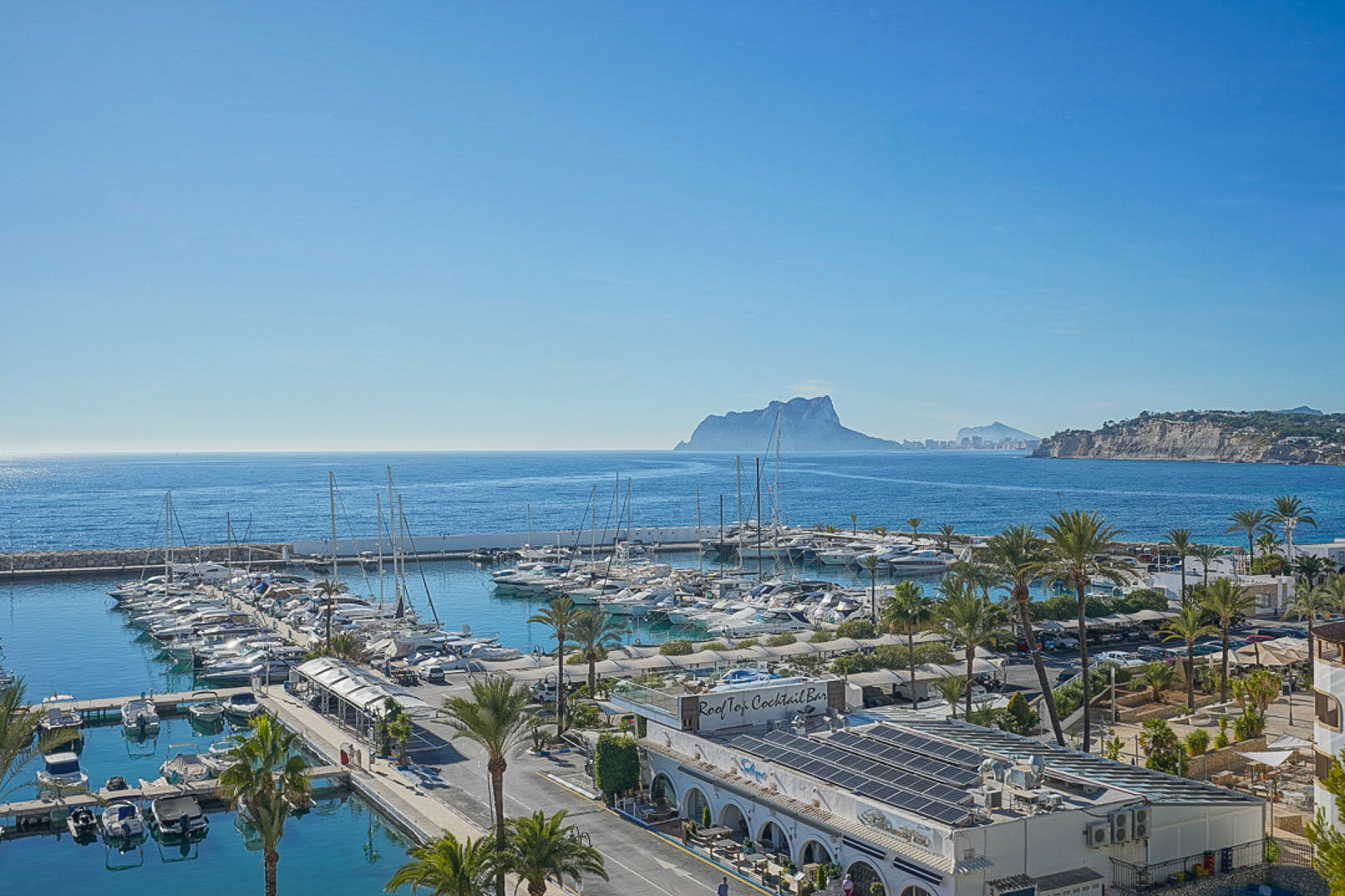 Panoramablick auf den Yachthafen von Moraira mit zahlreichen Booten, Palmen und dem markanten Peñón de Ifach im Hintergrund unter blauem Himmel.