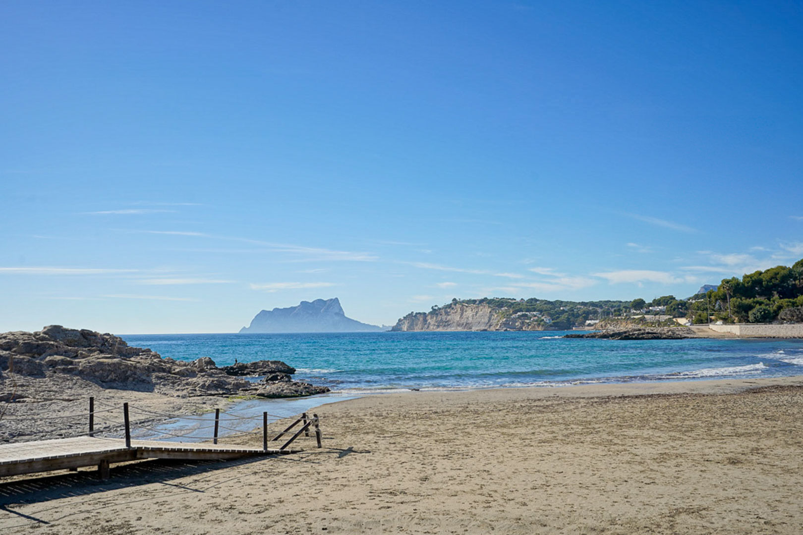 Sandstrand in Moraira mit Holzsteg, kristallklarem Mittelmeerwasser und weitem Blick auf den markanten Peñón de Ifach am Horizont.