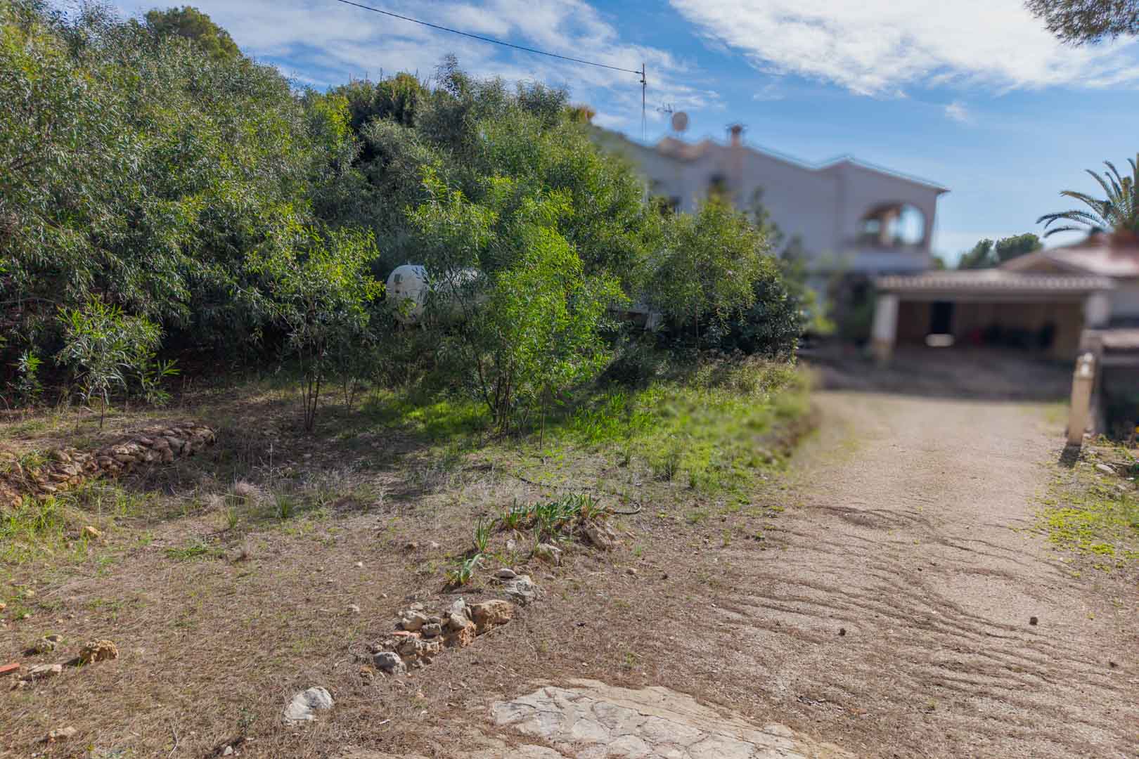 Zufahrtsweg zum Baugrundstück mit natürlichen Steinmauern und mediterranen Sträuchern, angrenzend an ein bestehendes Wohnhaus.