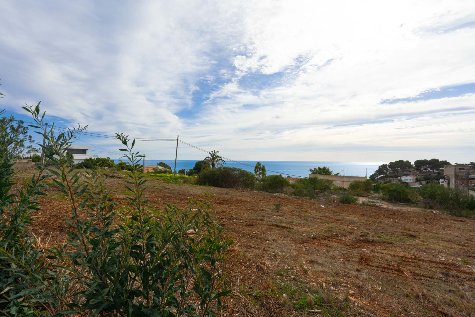 Erhöhtes Baugrundstück mit Panoramablick auf das Meer, geräumter Erdboden und mediterrane Vegetation unter leicht bewölktem Himmel.