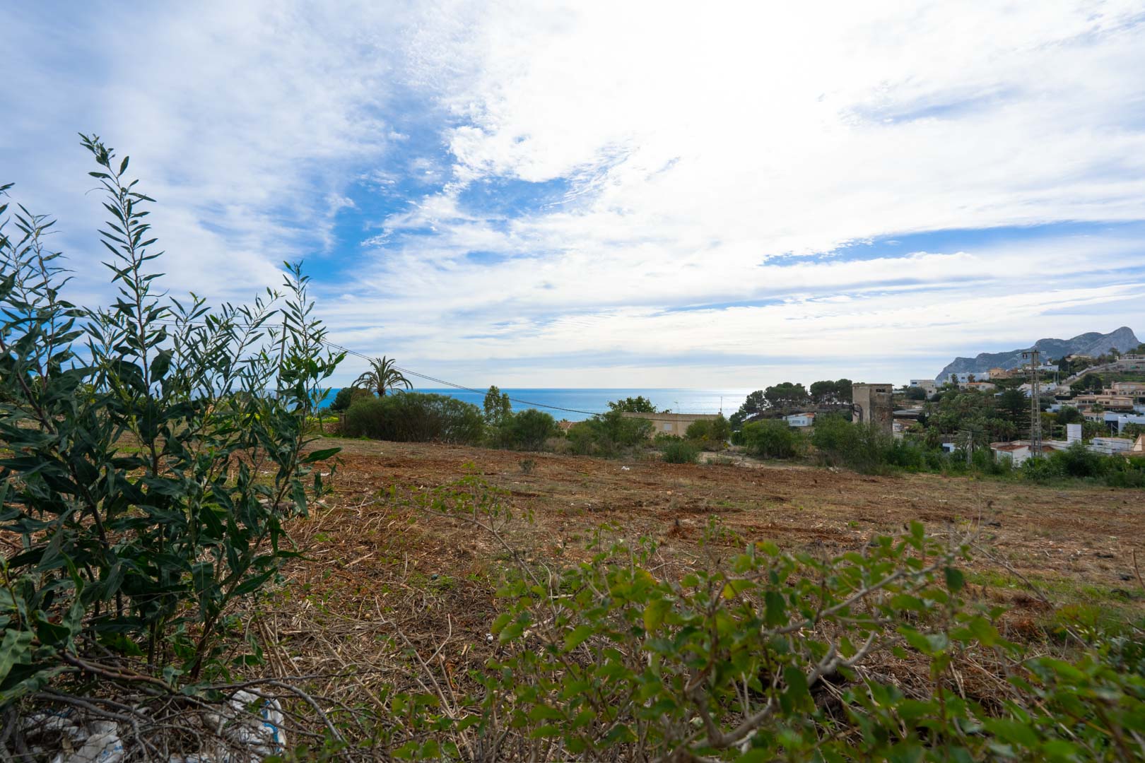 Erhöhtes Baugrundstück mit Panoramablick auf das Mittelmeer und die Berge, eingebettet in eine küstennahe Wohngegend mit natürlicher Vegetation.