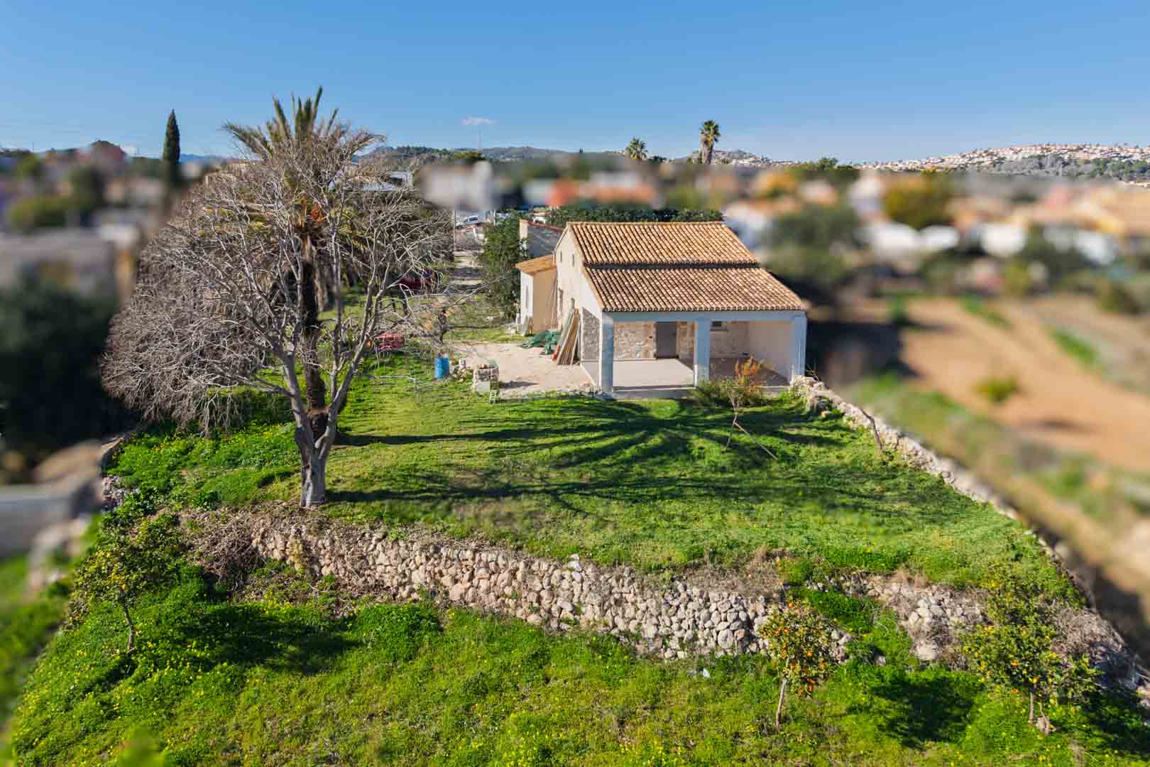 Luftaufnahme der traditionellen Finca mit renoviertem Ziegeldach, Trockensteinmauern und einer großen Naya, umgeben von einem grünen Grundstück mit Obstbäumen.