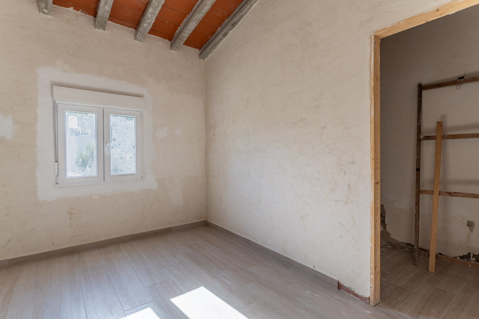 Renovierter Raum mit Bodenfliesen in Holzoptik, weißem PVC-Fenster und einer Dachschräge mit sichtbaren Betonbalken und Terrakotta-Elementen.