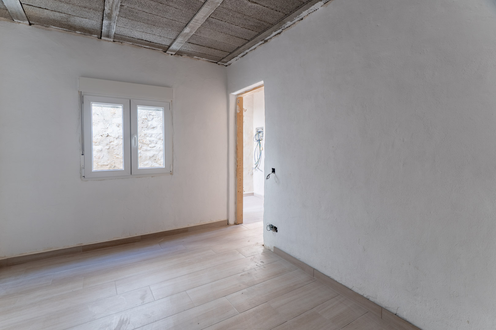 Renovierter Raum mit Bodenfliesen in Holzoptik, weißen Wänden und neuem PVC-Fenster. Die Decke zeigt die konstruktiven Betonbalken der Struktur.