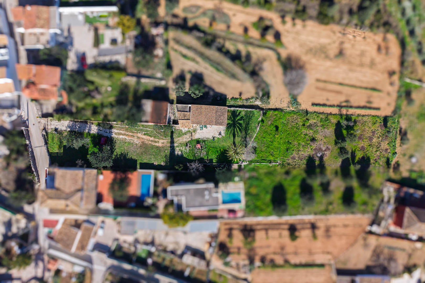 Zenitale Luftaufnahme des Anwesens und seines rechteckigen Grundstücks, die das zentrale Haus mit Ziegeldach und Gartenbereiche mit Palmen zeigt.