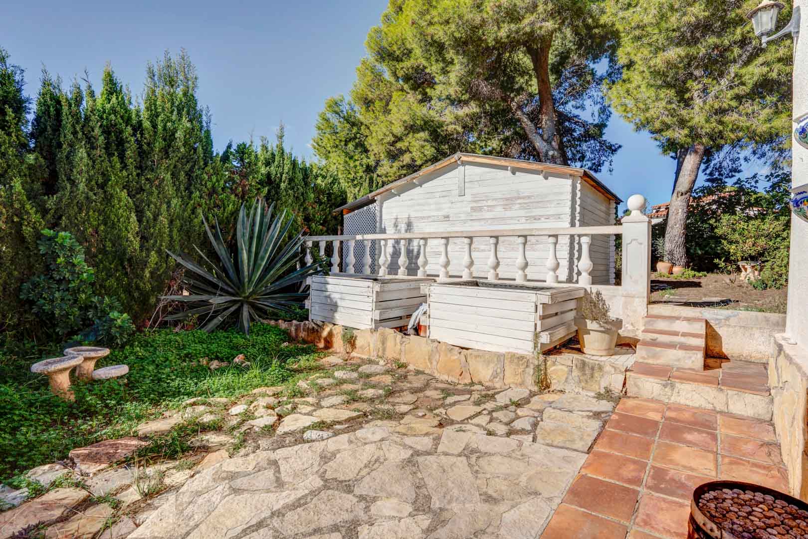 Außenbereich mit unregelmäßiger Natursteinpflasterung, weißem Holzschuppen und mediterraner Bepflanzung aus Pinien und einer großen Agave.
