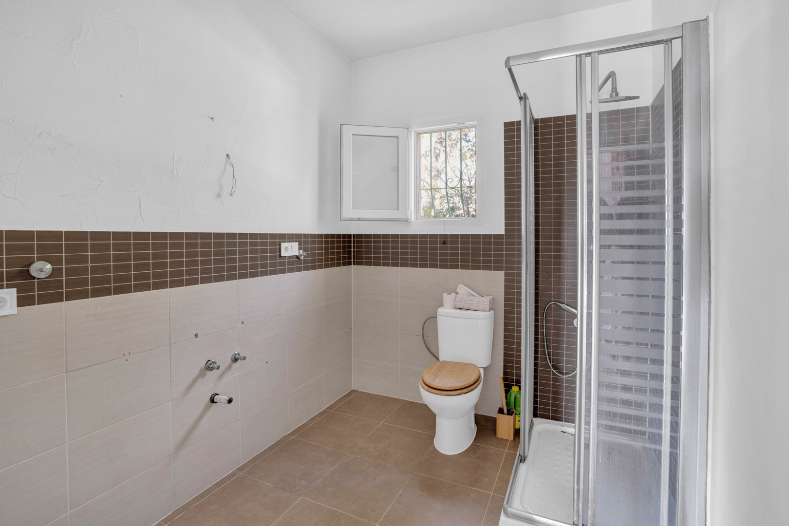 Badezimmer mit begehbarer Dusche, braunen Mosaikfliesen und einem WC mit Holzdeckel. Die Wände sind in neutralen Tönen gefliest und ein kleines Fenster sorgt für Tageslicht.