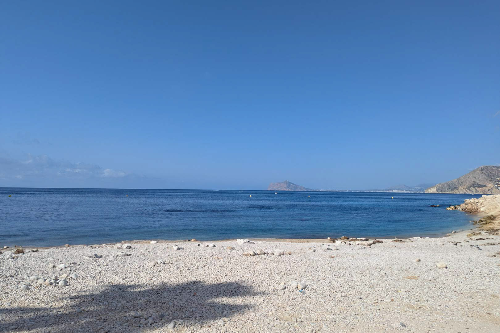 Blick auf eine weiße Kieselbucht mit ruhigem blauem Wasser in Calpe, mit der mediterranen Küstenlinie und fernen Bergen unter klarem Himmel.