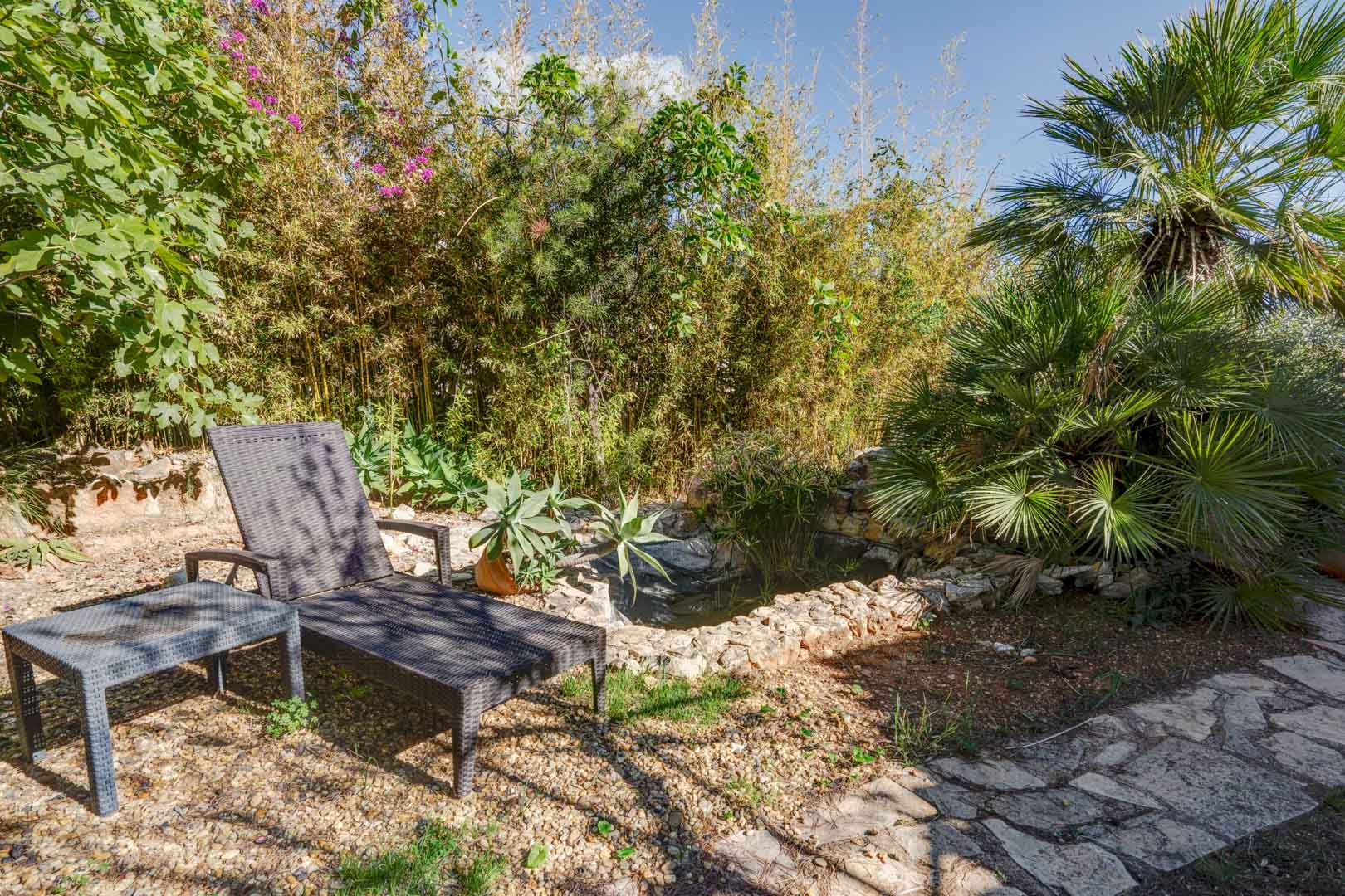 Entspannungsbereich im Garten mit Rattan-Sonnenliege neben einem Natursteinteich, umgeben von mediterraner Bepflanzung wie Bambus und Palmen.