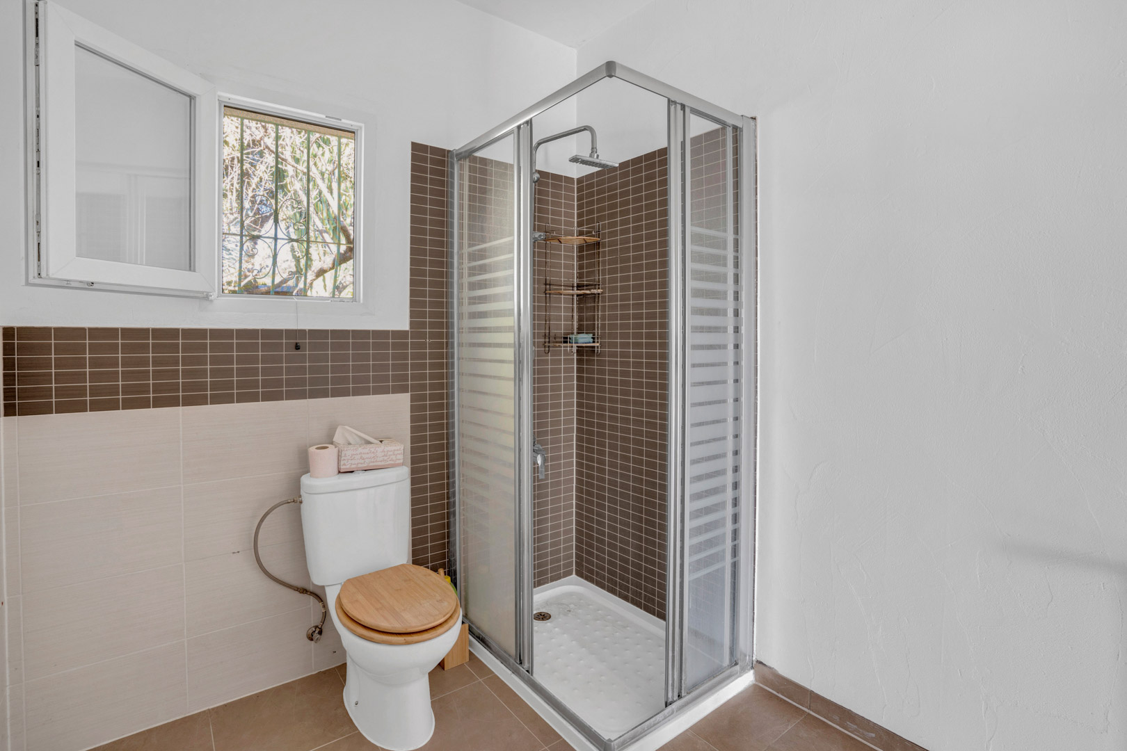 Funktionales Badezimmer mit Eckdusche, braunen Mosaikfliesen, Glasabtrennung, WC mit Holzdeckel und Fenster für natürliches Licht und Belüftung.