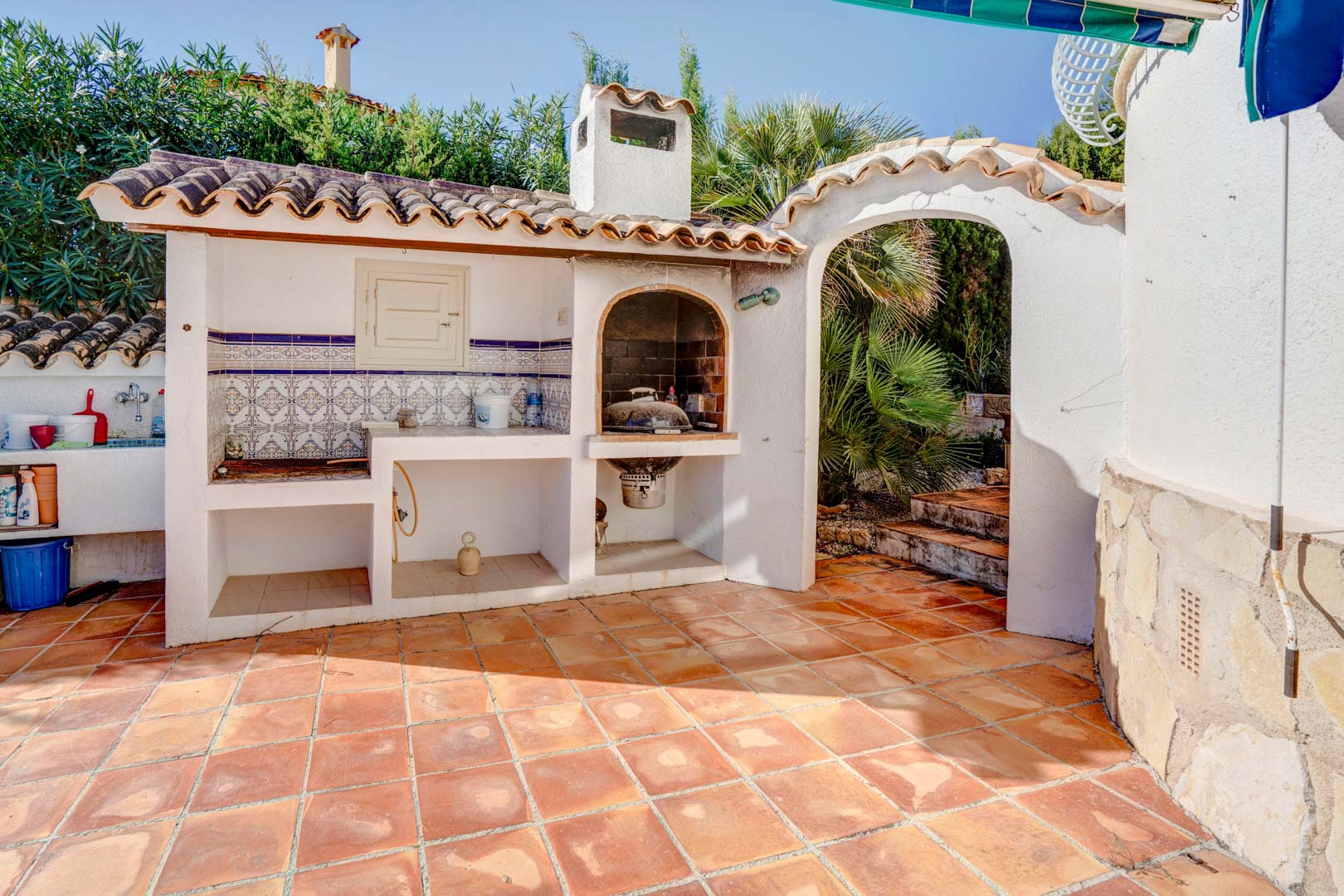 Gemauerter Grillbereich mit Kamin, Arbeitsplatte mit traditionellen Fliesen und Terrakottaboden, direkt neben einem Rundbogenzugang zum Garten.