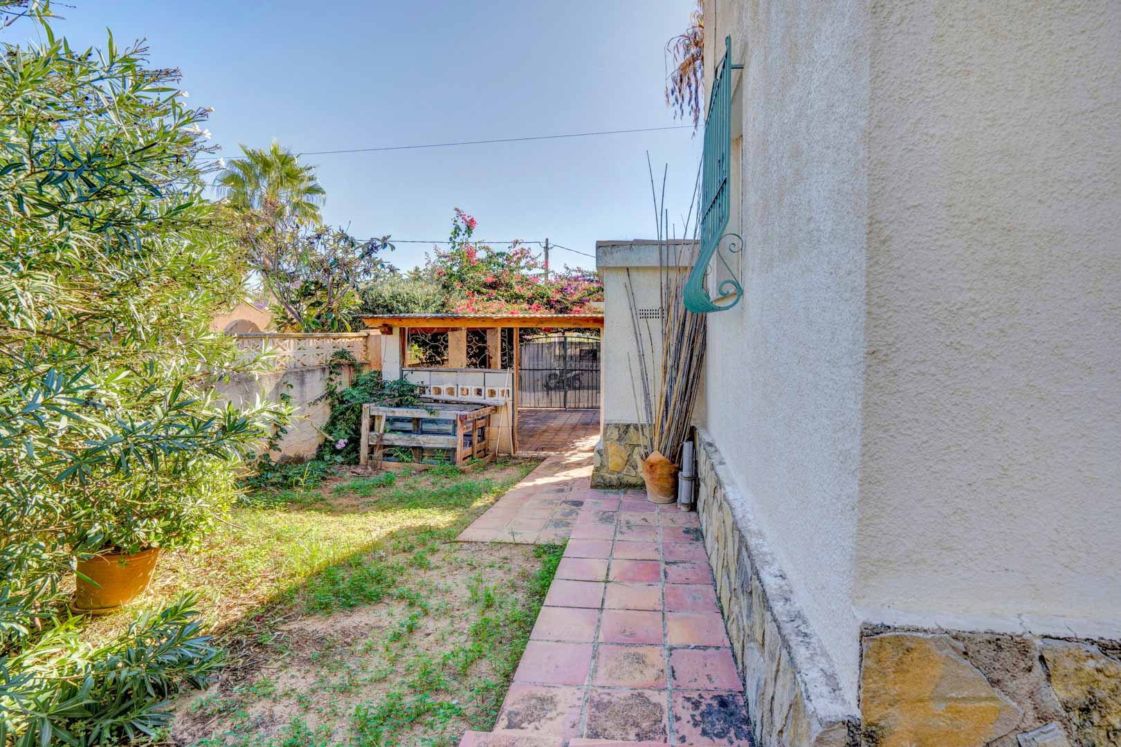 Gepflasterter Seitenweg zum überdachten Carport, mit weißen Putzwänden, Natursteinsockel und mediterraner Bepflanzung im Außenbereich.