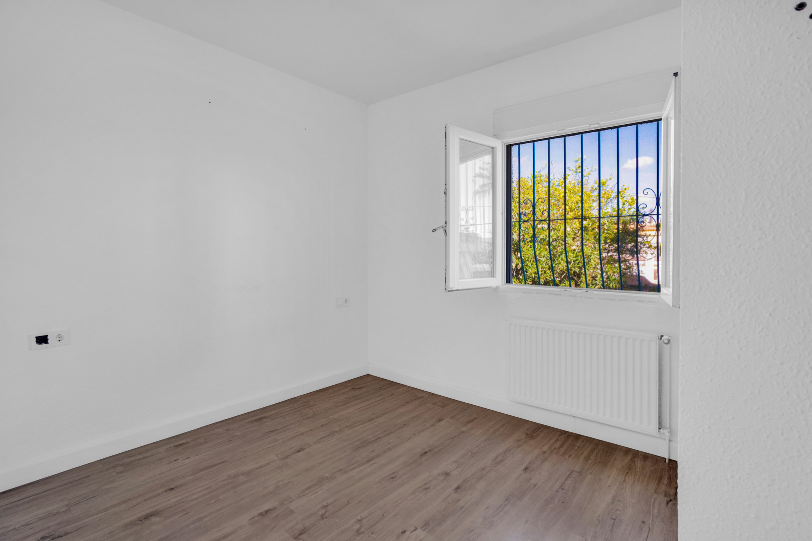 Heller Raum mit Laminatboden in Holzoptik, weißen Wänden und Fenster mit dekorativen Gittern, Gartenblick und installiertem Heizkörper.