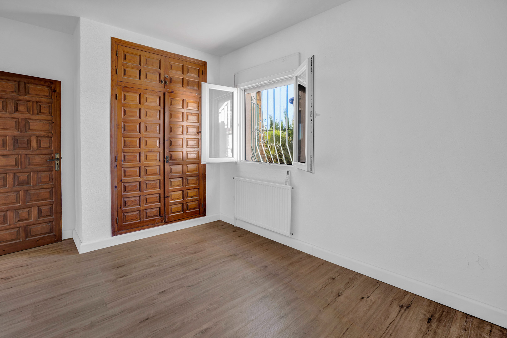 Helles Schlafzimmer mit Einbauschrank aus geschnitztem Holz, Laminatboden, Heizkörper und weißem PVC-Fenster mit Blick in den Garten.