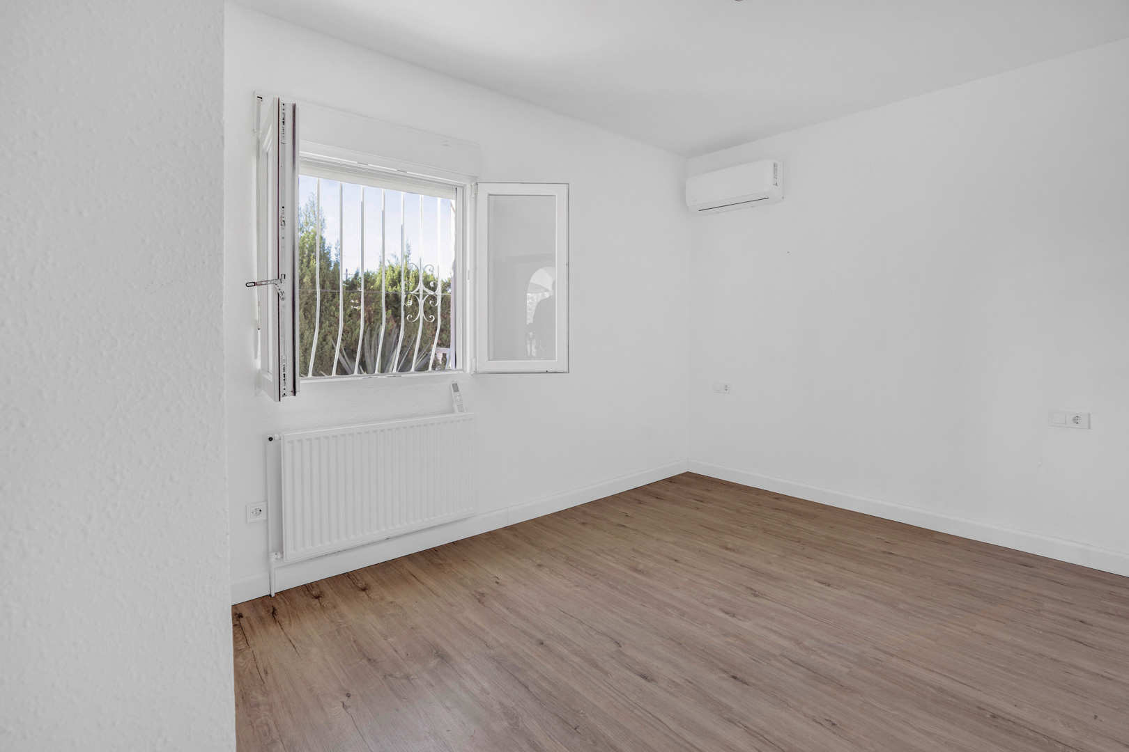 Helles Schlafzimmer mit Laminatboden in Holzoptik, ausgestattet mit einem Zentralheizungskörper und einer Split-Klimaanlage an weißen Wänden.