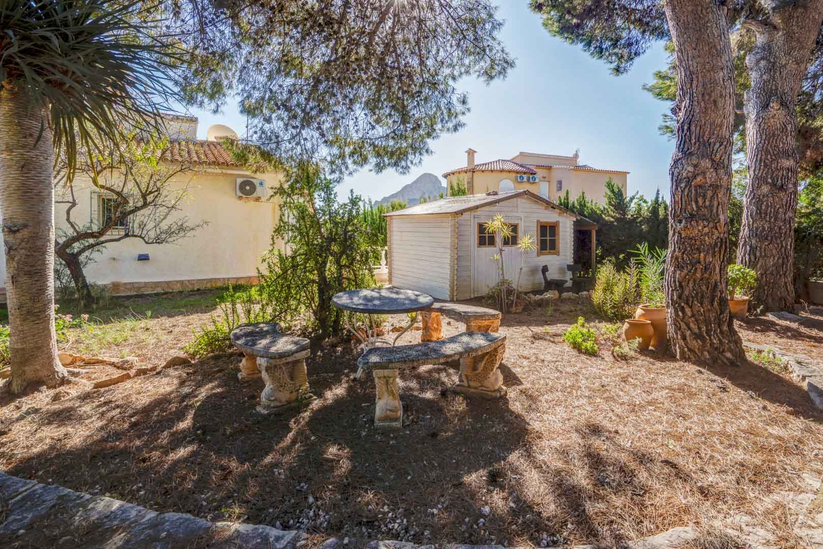 Mediterraner Garten mit Steinsitzgruppe unter Pinien, weißem Holzgartenhaus und Fernblick auf den Peñón de Ifach.
