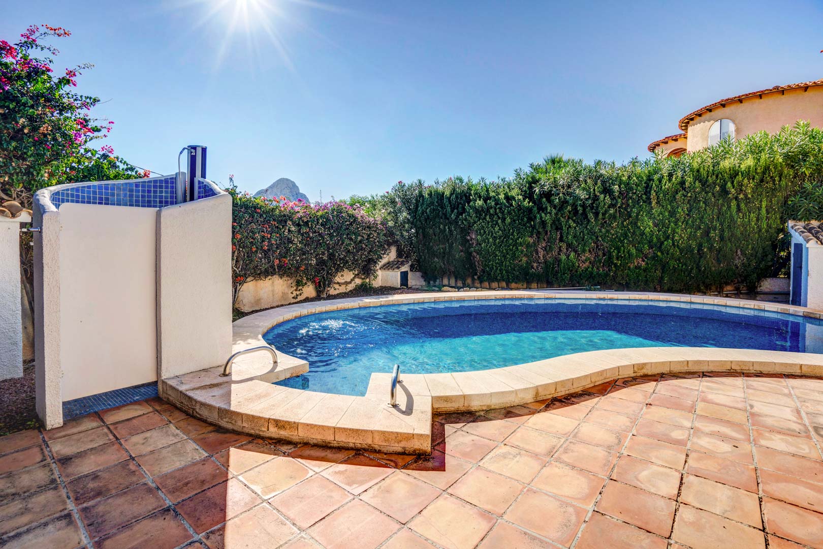 Nierenförmiger Privatpool mit Terrakotta-Terrasse, Außendusche und Blick auf den Peñón de Ifach hinter hohen Hecken, die für Privatsphäre sorgen.