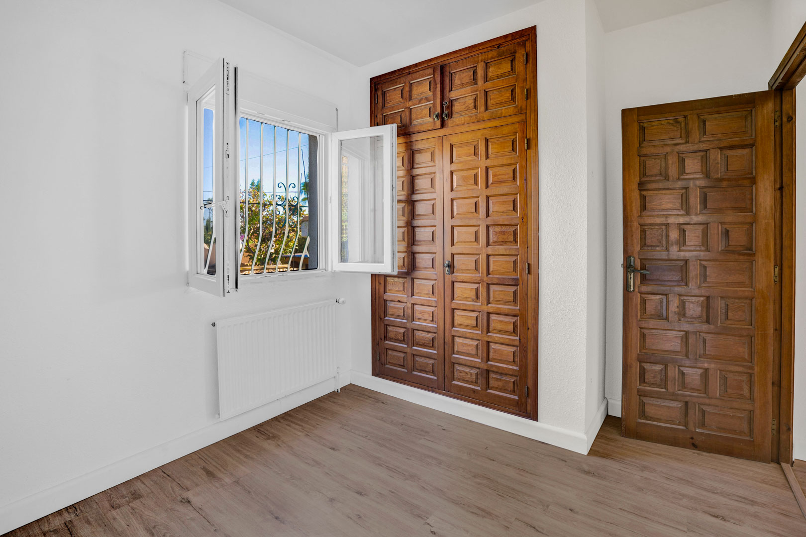 Schlafzimmer mit eingebautem Holzschrank mit Kassettenfronten, Laminatboden, Zentralheizungskörper und Fenster mit natürlichem Lichteinfall.