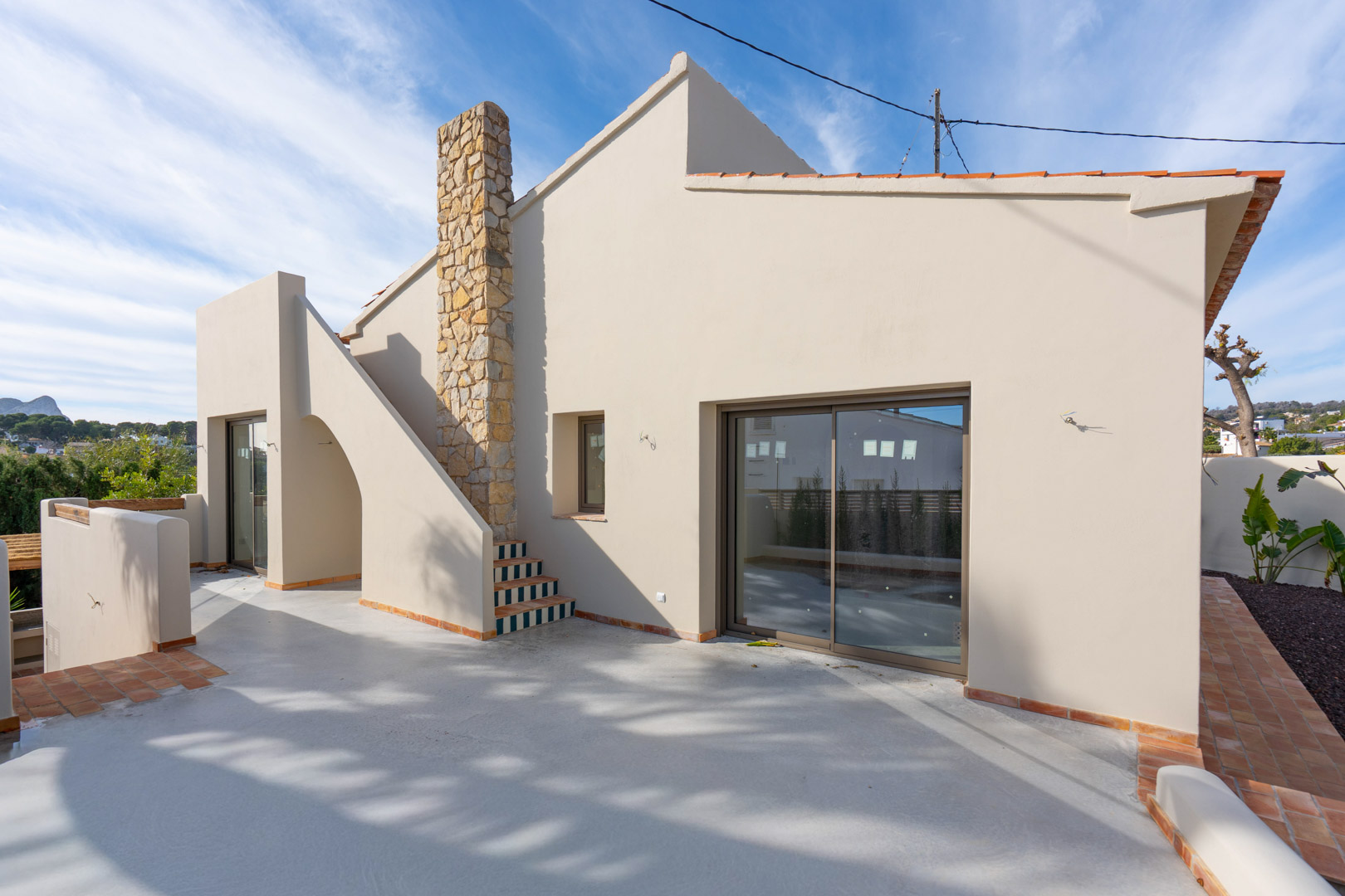 Außenansicht der Villa mit polierter Beton-Terrasse, Natursteinkamin und dekorativer Treppe. Im Hintergrund ist der Peñón de Ifach zu sehen.