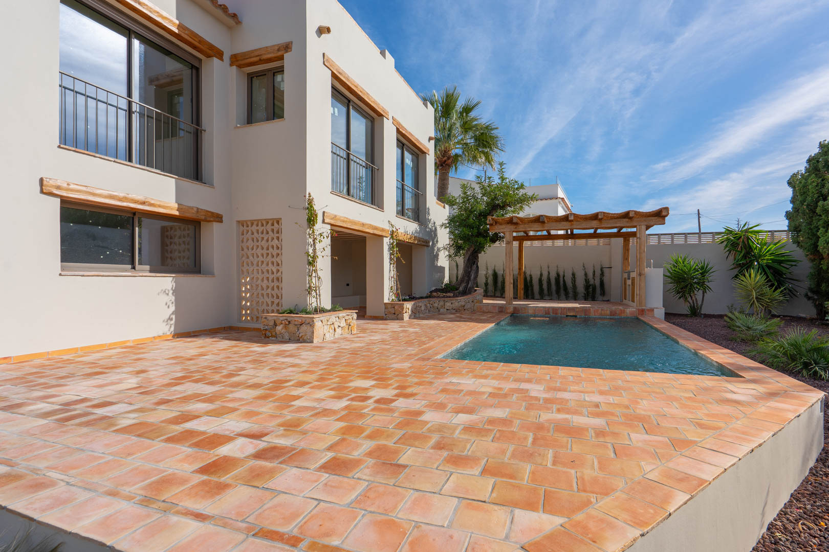 Außenansicht mit großer Terrakotta-Terrasse, neuem Pool und Holzpergola. Die Fassade besticht durch Fensterstürze aus Naturholz und integrierte Steinbeete.