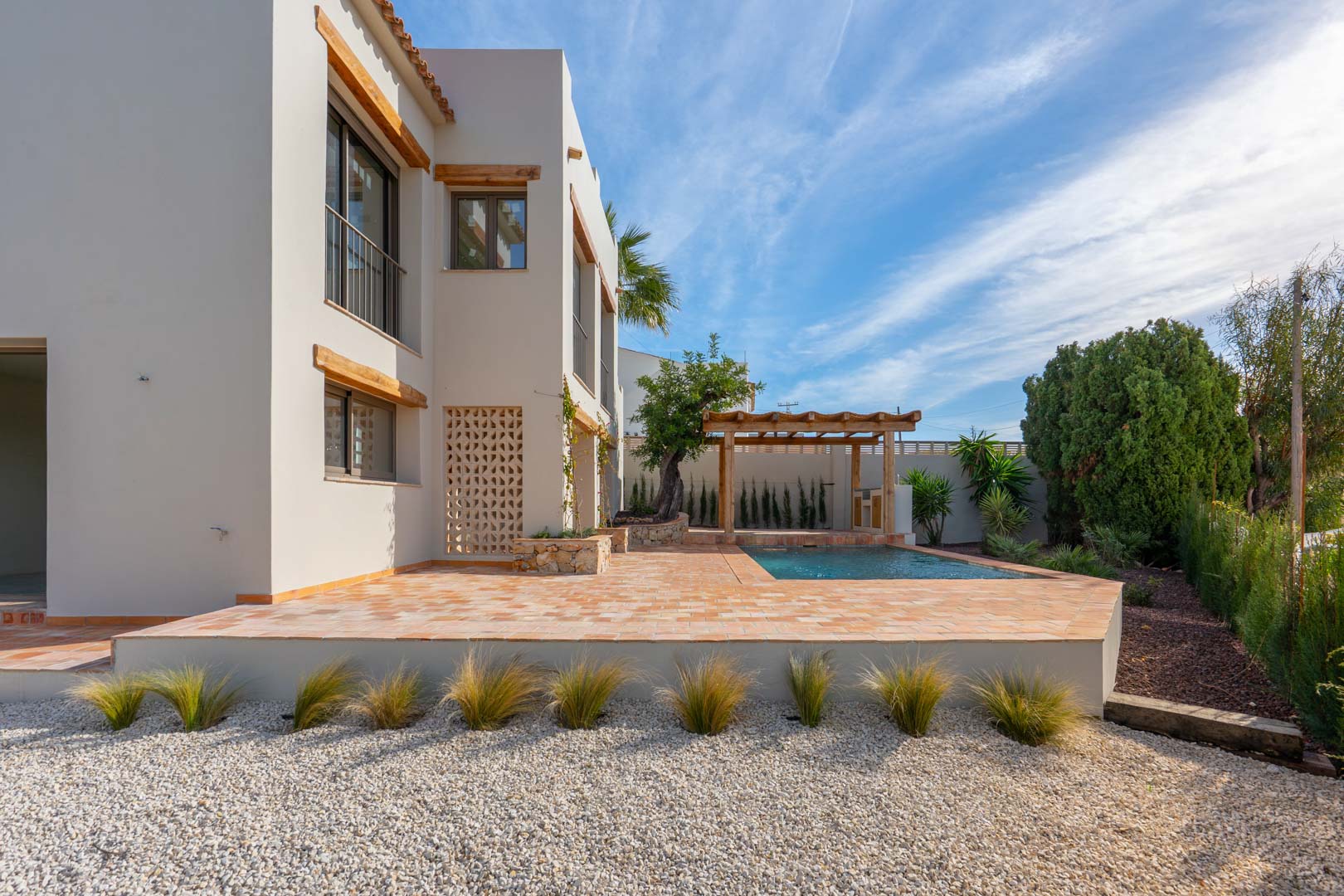 Außenansicht mit Terrakotta-Terrasse, neu gebautem Pool und Holzpergola in einem mediterran angelegten Garten mit Kies und Ziergräsern.