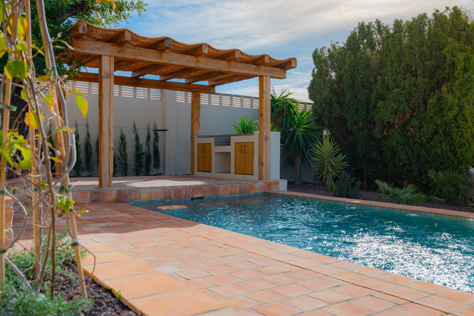 Außenbereich mit blau gefliestem Pool, Terrakotta-Boden und einer Holzpergola mit integrierter Außenküche, umgeben von mediterraner Bepflanzung.
