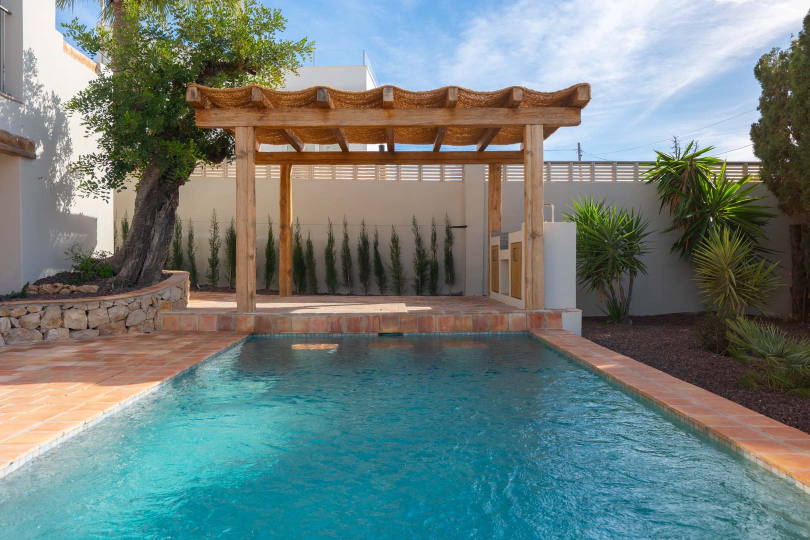 Außenpool mit Terrakotta-Fliesen und einer rustikalen Holzpergola samt Grillstation, eingebettet in einen mediterranen Garten mit Olivenbäumen und Zypressen.