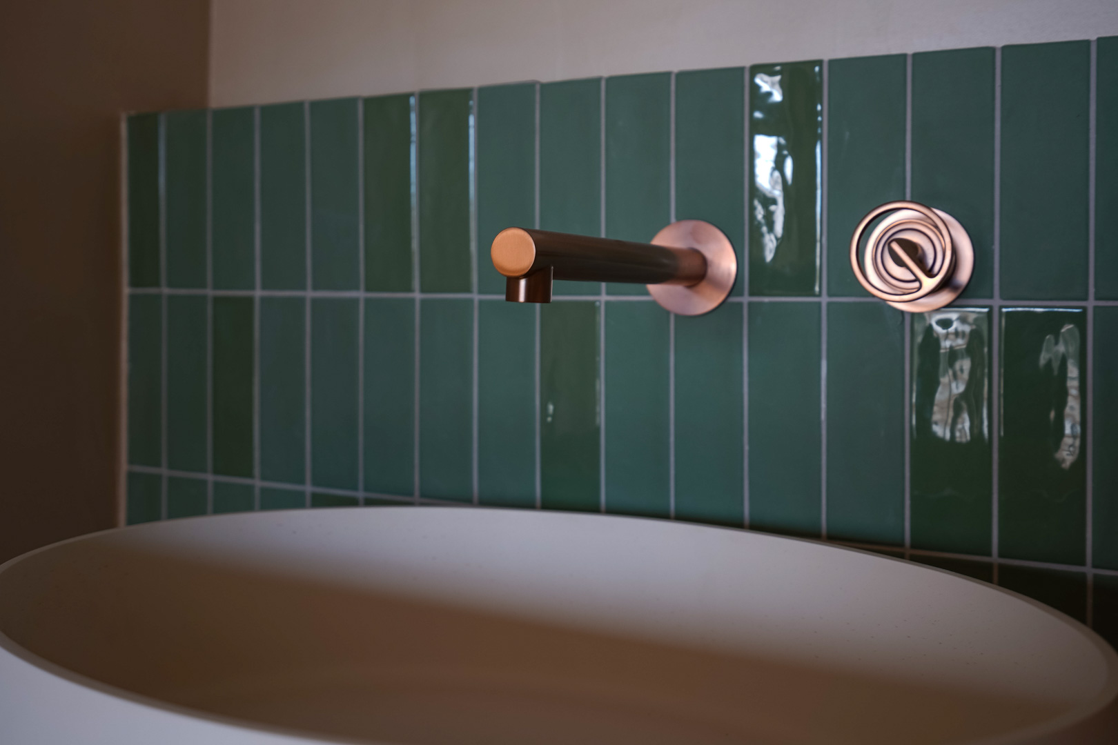Badezimmerdetail mit wandmontierter Kupferarmatur, glänzenden vertikalen Fliesen in Smaragdgrün und einem mattweißen Aufsatzwaschbecken.