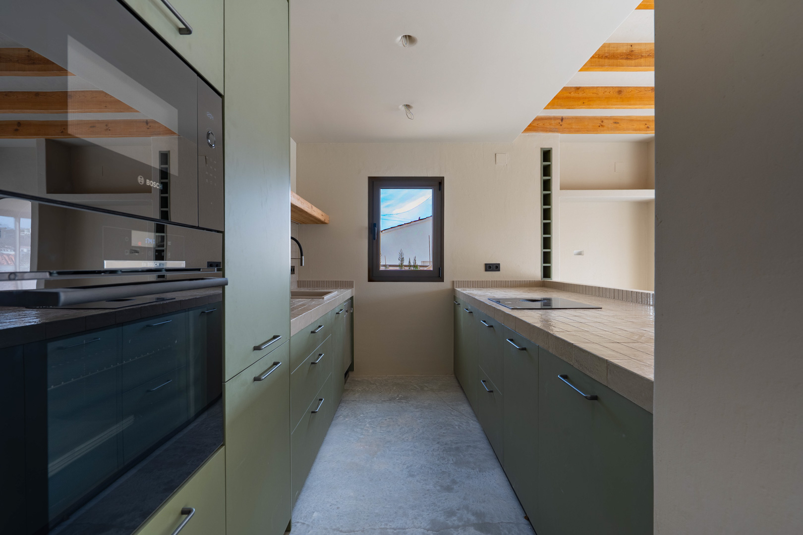 Designerküche mit olivgrünen Schränken, integrierten Bosch-Geräten und gefliesten Arbeitsplatten. Sichtbare Holzbalken und ein polierter Betonboden prägen den Raum.