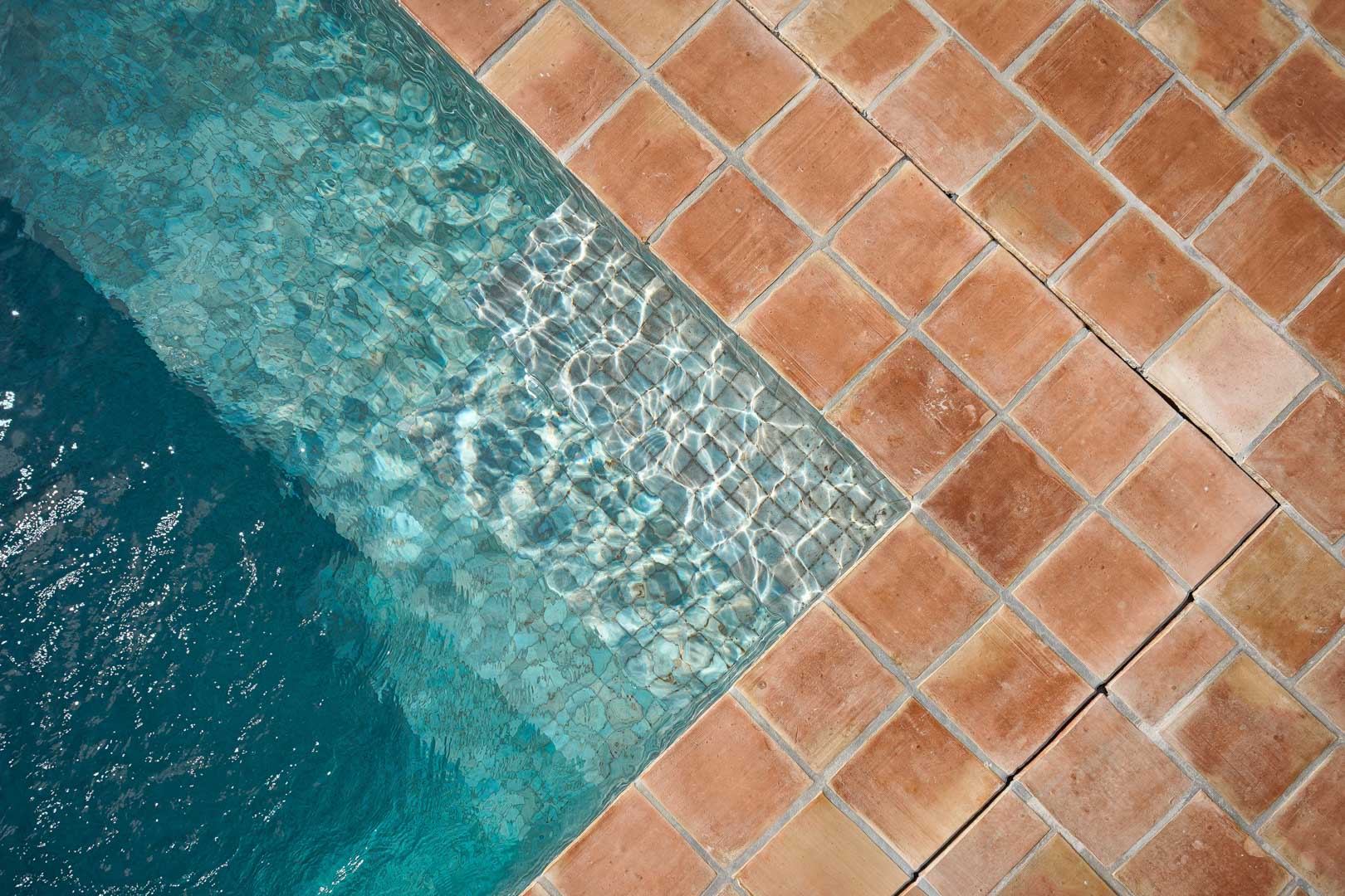 Detailaufnahme der Poolecke mit handgefertigten Terrakotta-Fliesen, die einen warmen Kontrast zum klaren, türkisfarbenen Wasser und den Lichtreflexionen bilden.