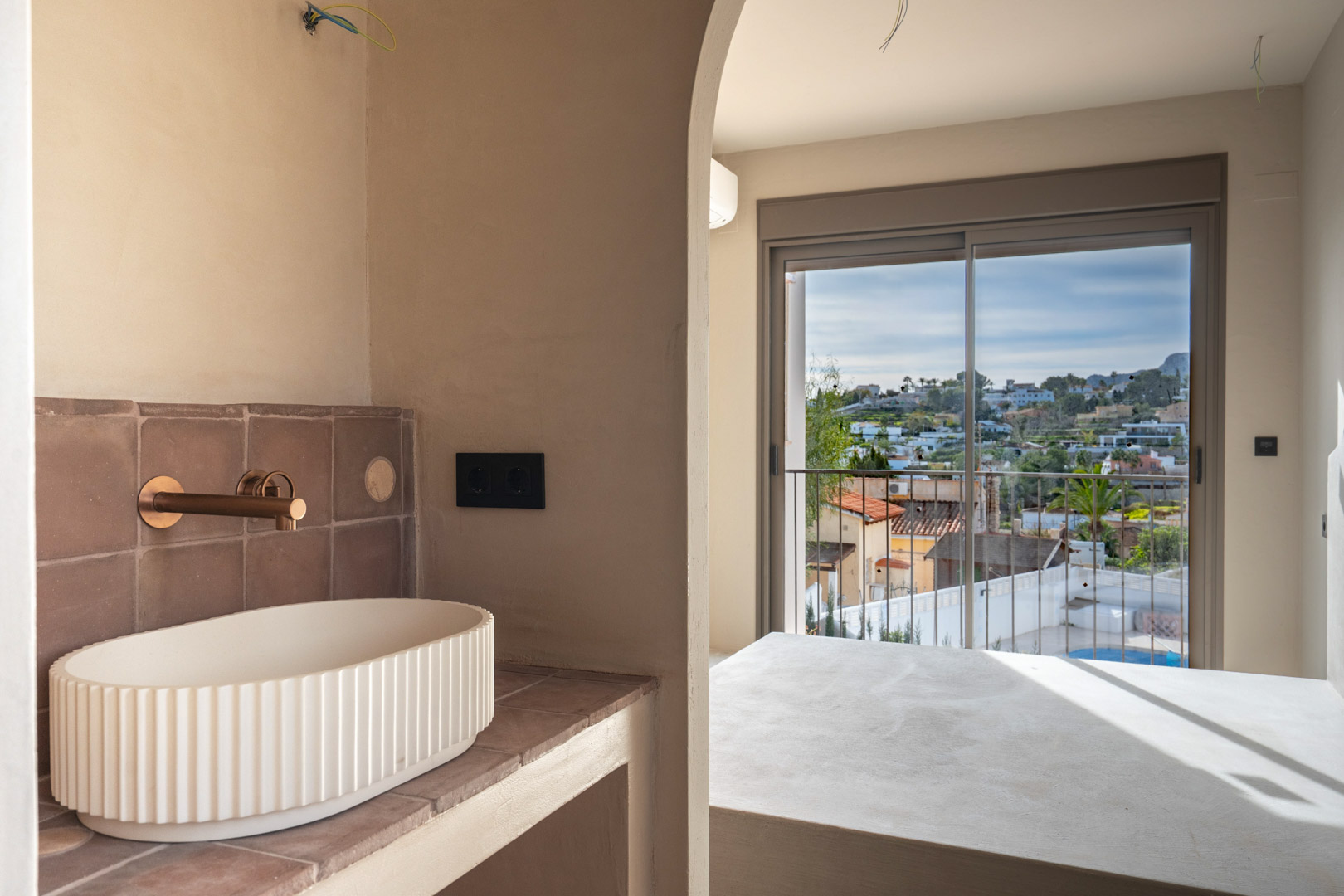 En-Suite-Badezimmer mit geriffeltem Waschbecken und Kupferarmaturen, das durch einen Rundbogen den Blick auf das Schlafzimmer und den Balkon freigibt.