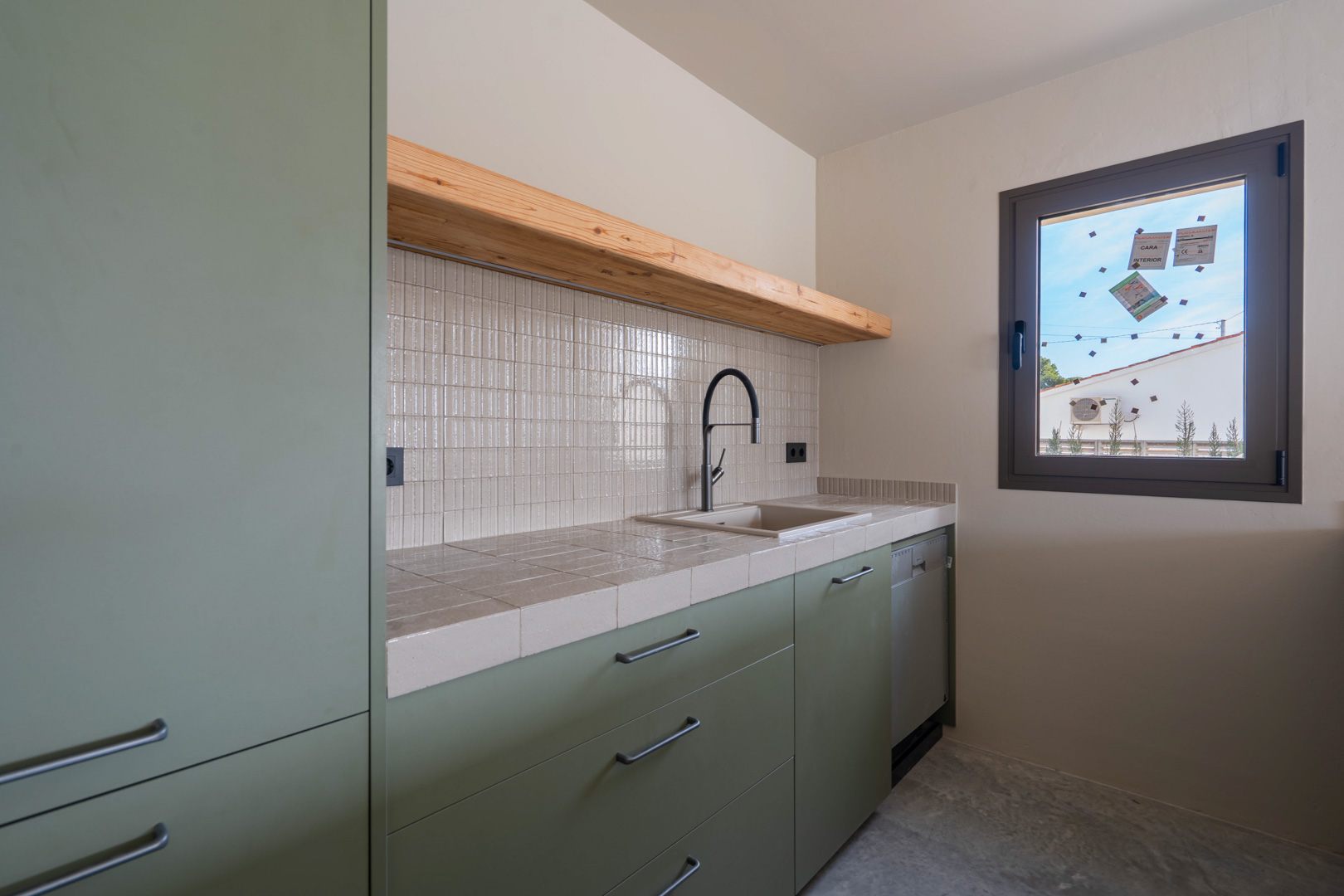 Gästeküche mit salbeigrünen Schränken, gefliester Arbeitsplatte, Massivholzregal und schwarzer Armatur auf poliertem Betonboden.