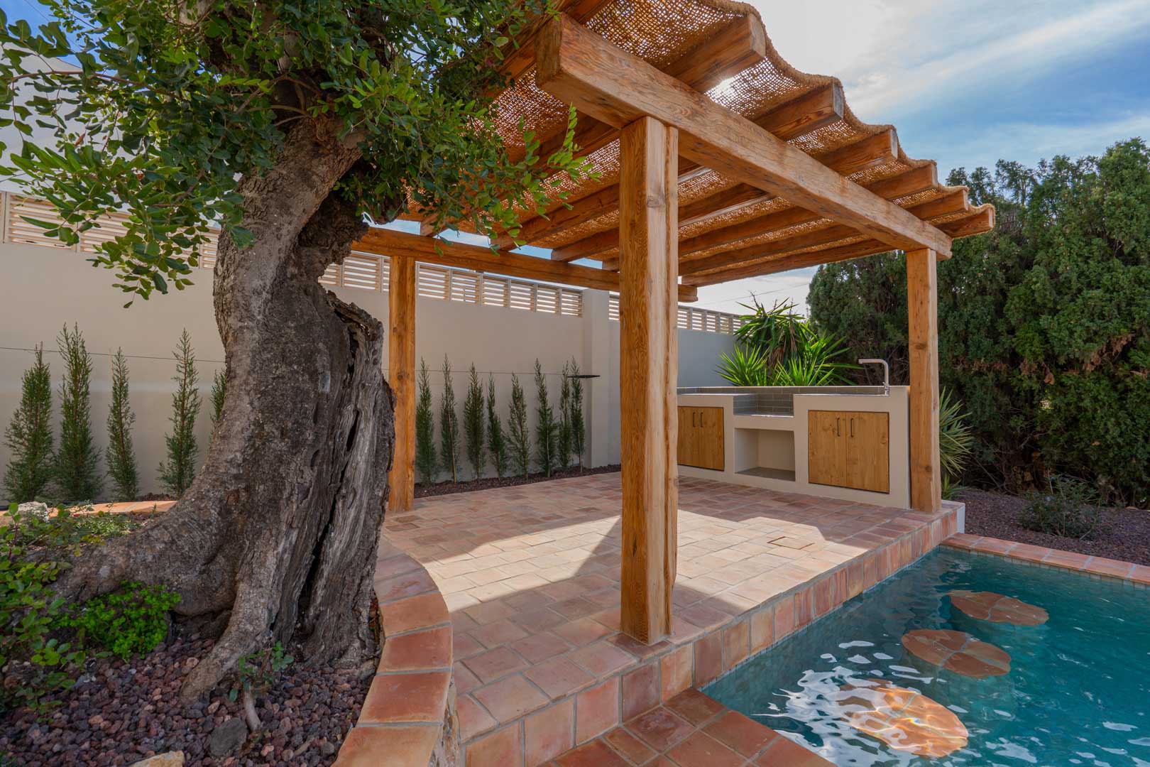 Holzpergola mit Naturfaserdach auf Terrakottaboden, ausgestattet mit Außenküche und einem Pool mit integrierten Barhockern neben einem alten Olivenbaum.