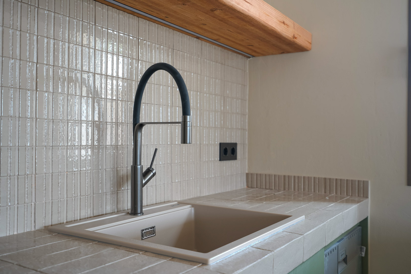 Küchendetail mit Arbeitsplatte aus Feinsteinzeugfliesen und integriertem Spülbecken. Moderne Flex-Armatur vor vertikalem Mosaik-Fliesenspiegel und Massivholzregal.