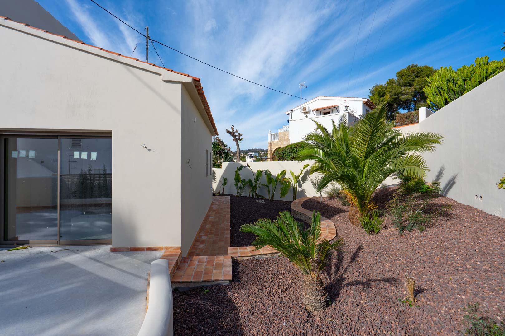 Mediterraner Garten mit Palmen und Vulkankies, eingefasst von Terrakotta-Pfaden und weißen Außenmauern.