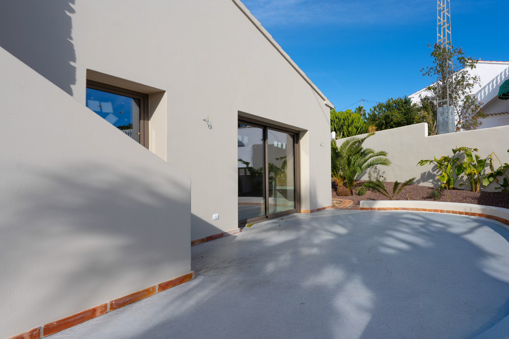 Minimalistische Fassade in Erdtönen mit großen Glasschiebetüren und einer geschwungenen Terrasse mit Terrakotta-Kanten und mediterraner Bepflanzung.