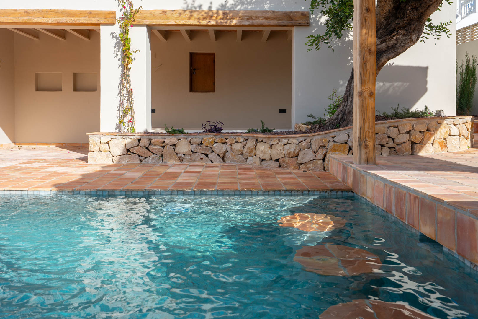 Neu gebauter Pool mit integrierten Unterwassersitzplätzen, umgeben von Terrakotta-Fliesen, Natursteinmauern und rustikalen Holzbalken im Außenbereich.