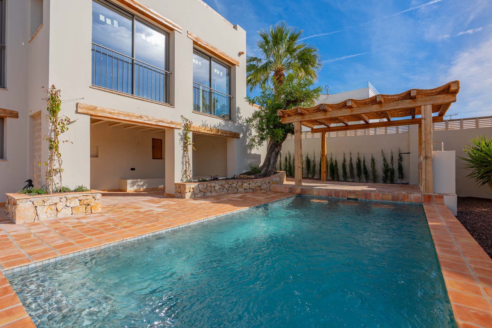 Neu gebauter Pool mit Terrakotta-Fliesen, einer Holzpergola und Natursteinmauern vor der renovierten Fassade mit mediterranen Holzelementen.