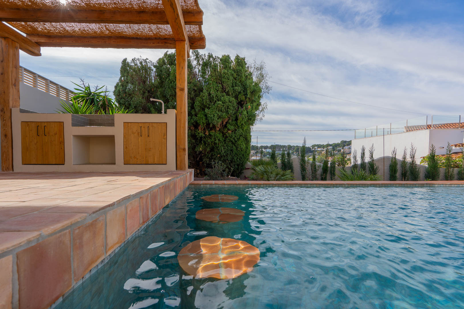 Poolbereich mit integrierten Unterwassersitzplätzen, Außenküche unter einer Holzpergola und Bodenbelag aus klassischen Terrakotta-Fliesen.
