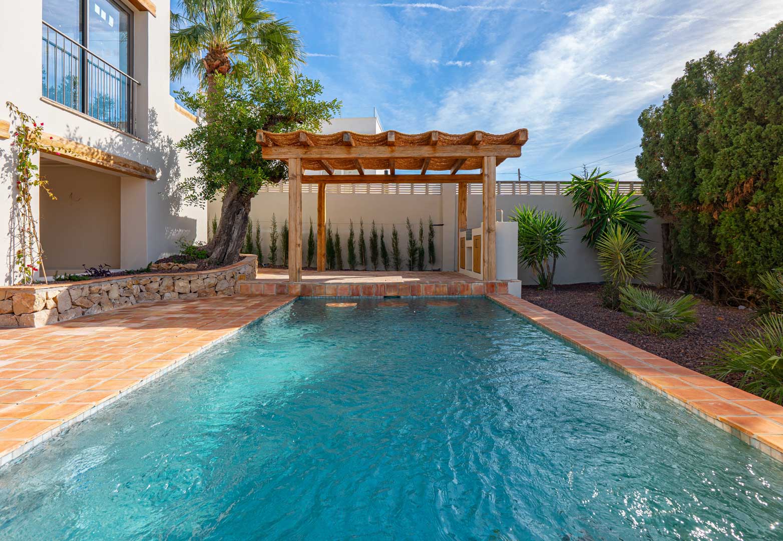 Rechteckiger Pool mit Terrakotta-Einfassung, Holzpergola und mediterranem Garten mit Natursteinmauern, Palmen und gepflegter Bepflanzung bei Tageslicht.
