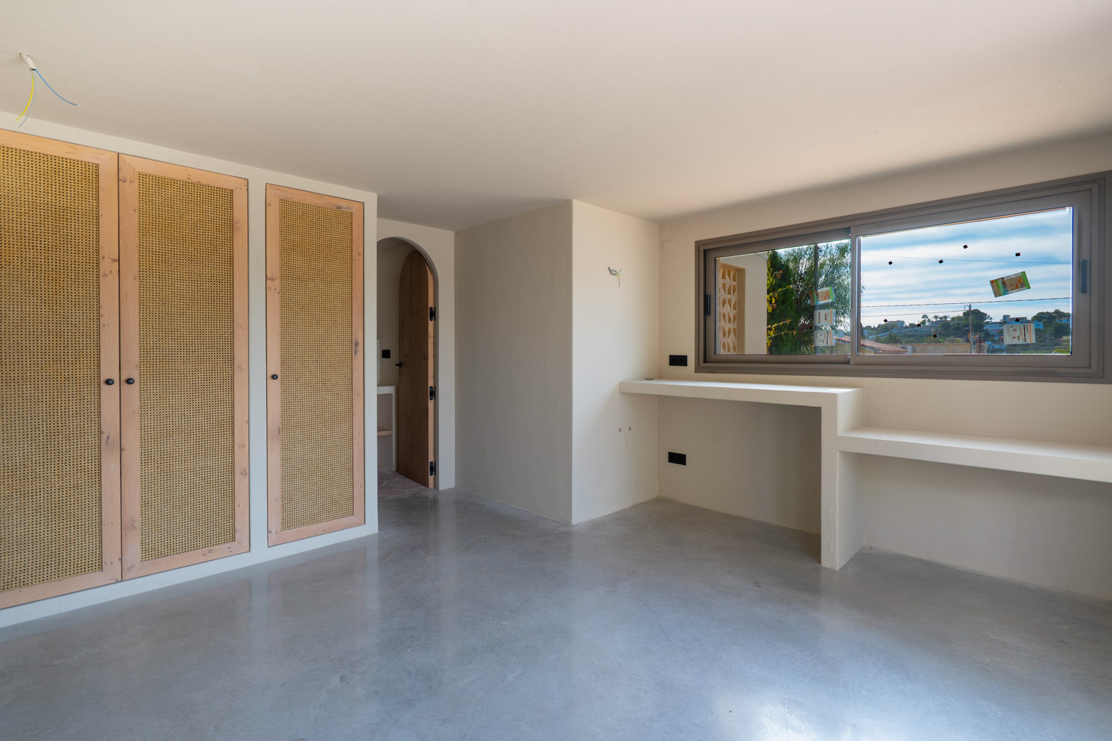 Schlafzimmer mit Einbauschränken aus Holz und Wiener Geflecht, poliertem Betonboden und integriertem Schreibtisch unter dem Fenster neben einem Rundbogen.