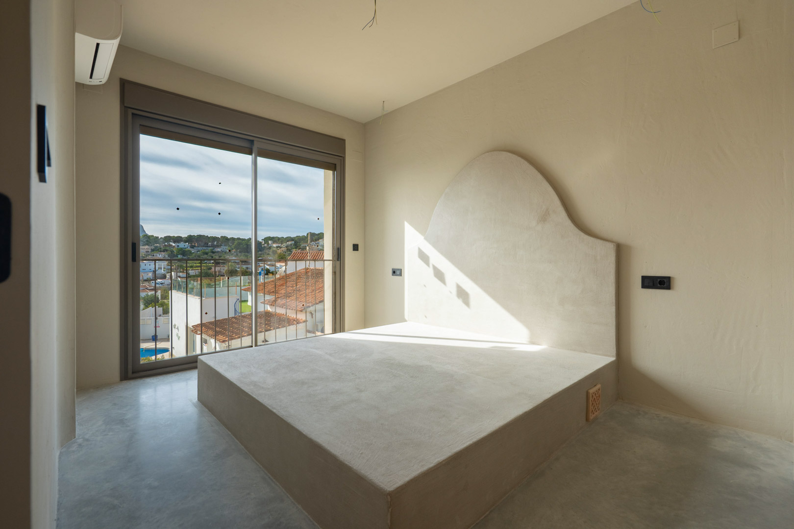 Schlafzimmer mit integriertem Bettgestell und geschwungenem Kopfteil aus Ton, poliertem Betonboden und Zugang zum Balkon mit Weitblick.