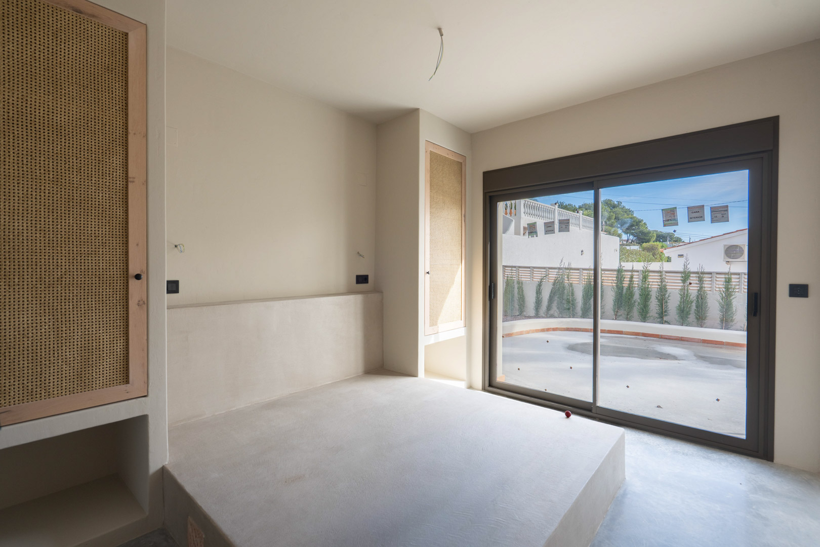 Schlafzimmer mit integriertem Bettsockel aus poliertem Beton, Einbauschränken mit Wiener Geflecht und direktem Zugang zur Terrasse durch Glasschiebetüren.