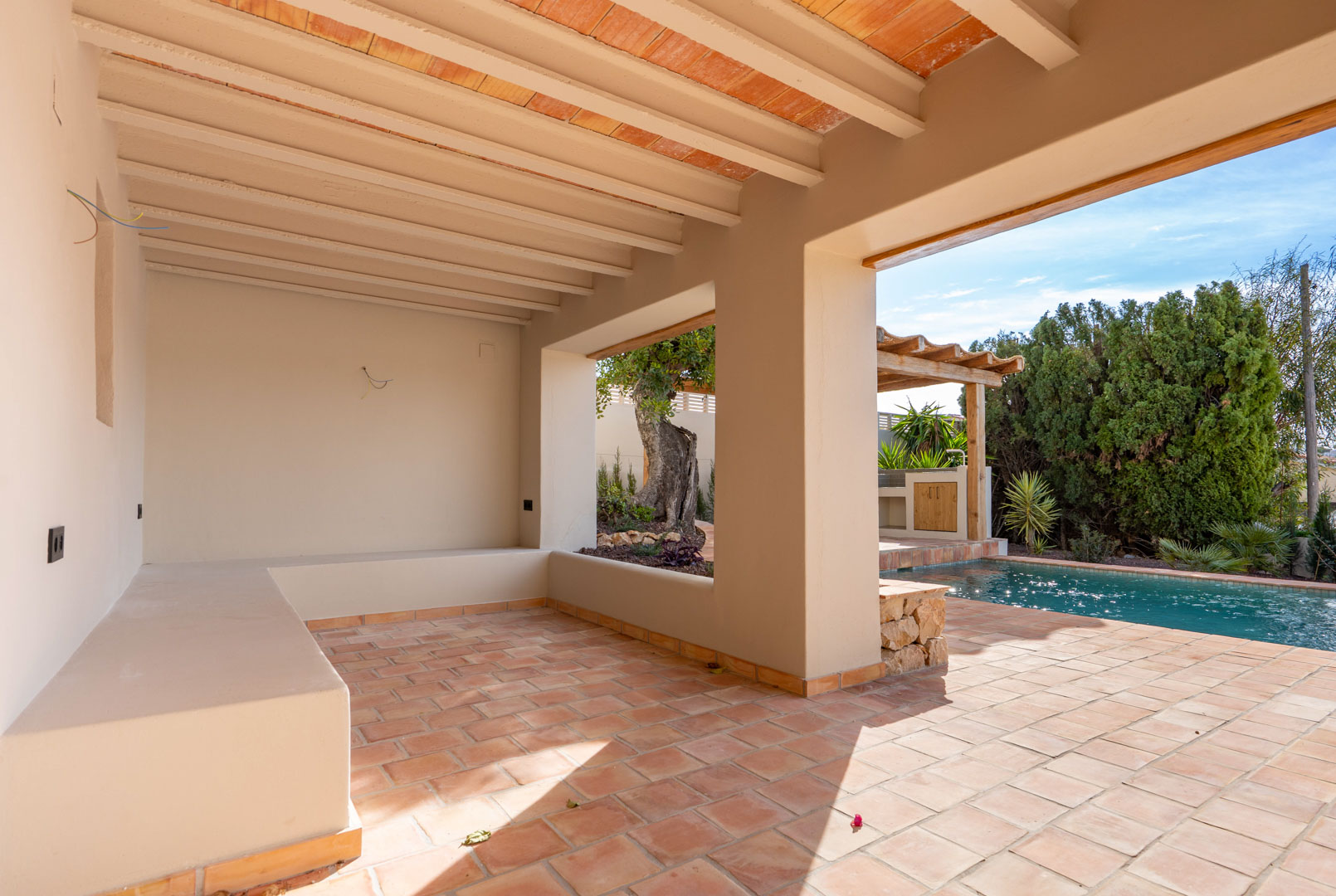 Überdachte Terrasse mit Terrakottaböden, Sichtbalkendecke und gemauerter Sitzbank mit Blick auf den Pool und die angrenzende Pergola.