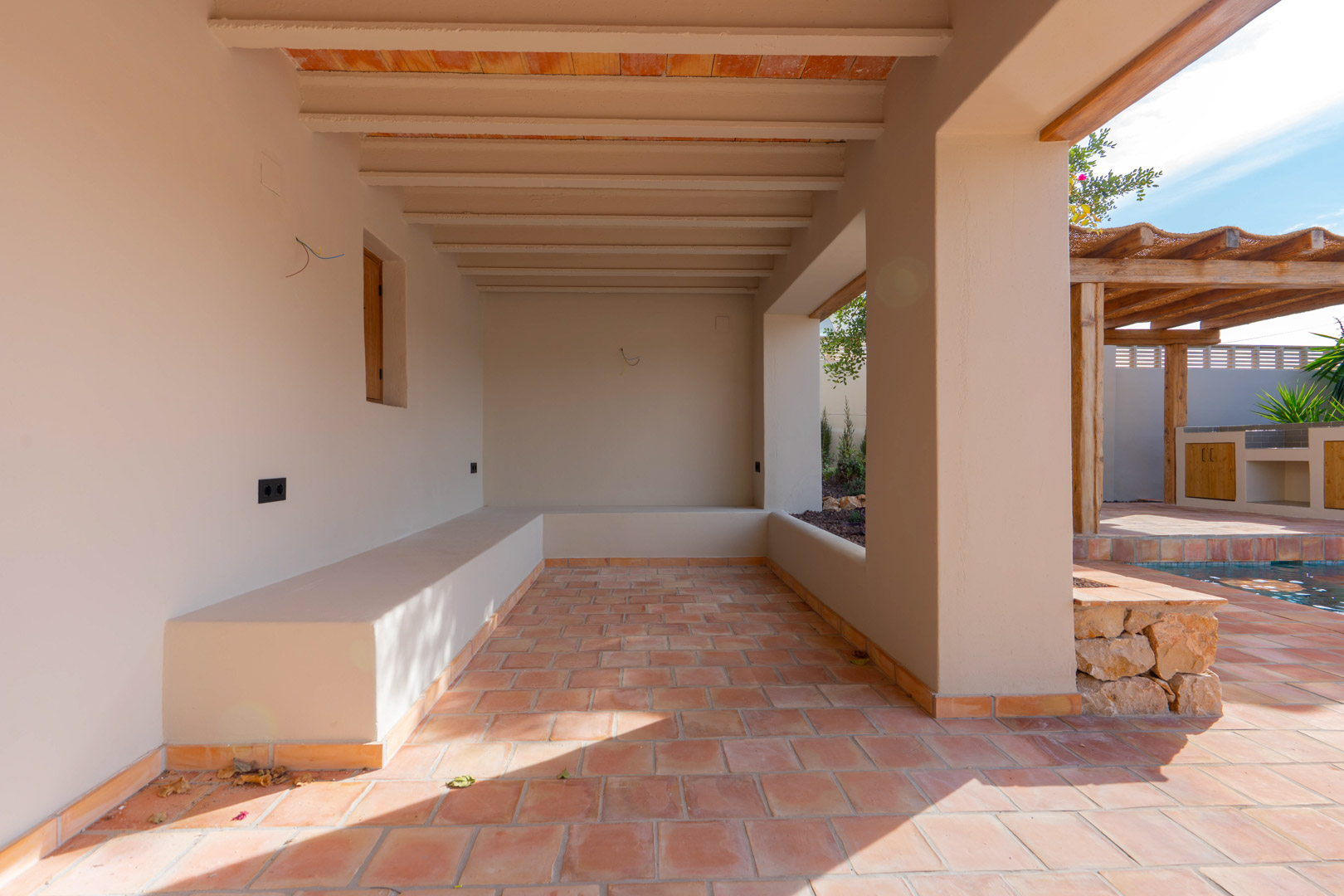 Überdachte Veranda mit Terrakottaboden, Sichtbalkendecke und integrierten Sitzbänken mit Blick auf den Poolbereich und die Pergola.