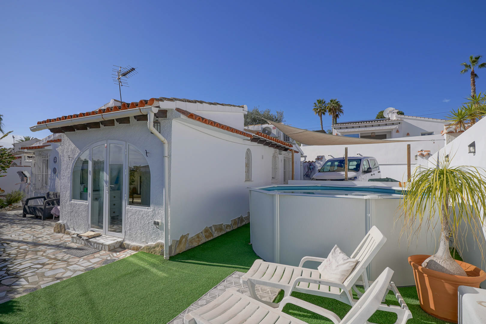 Außenansicht der weißen Villa mit verglaster Veranda, einem Aufstellpool auf Kunstrasen und einem Liegebereich mit weißen Sonnenstühlen.