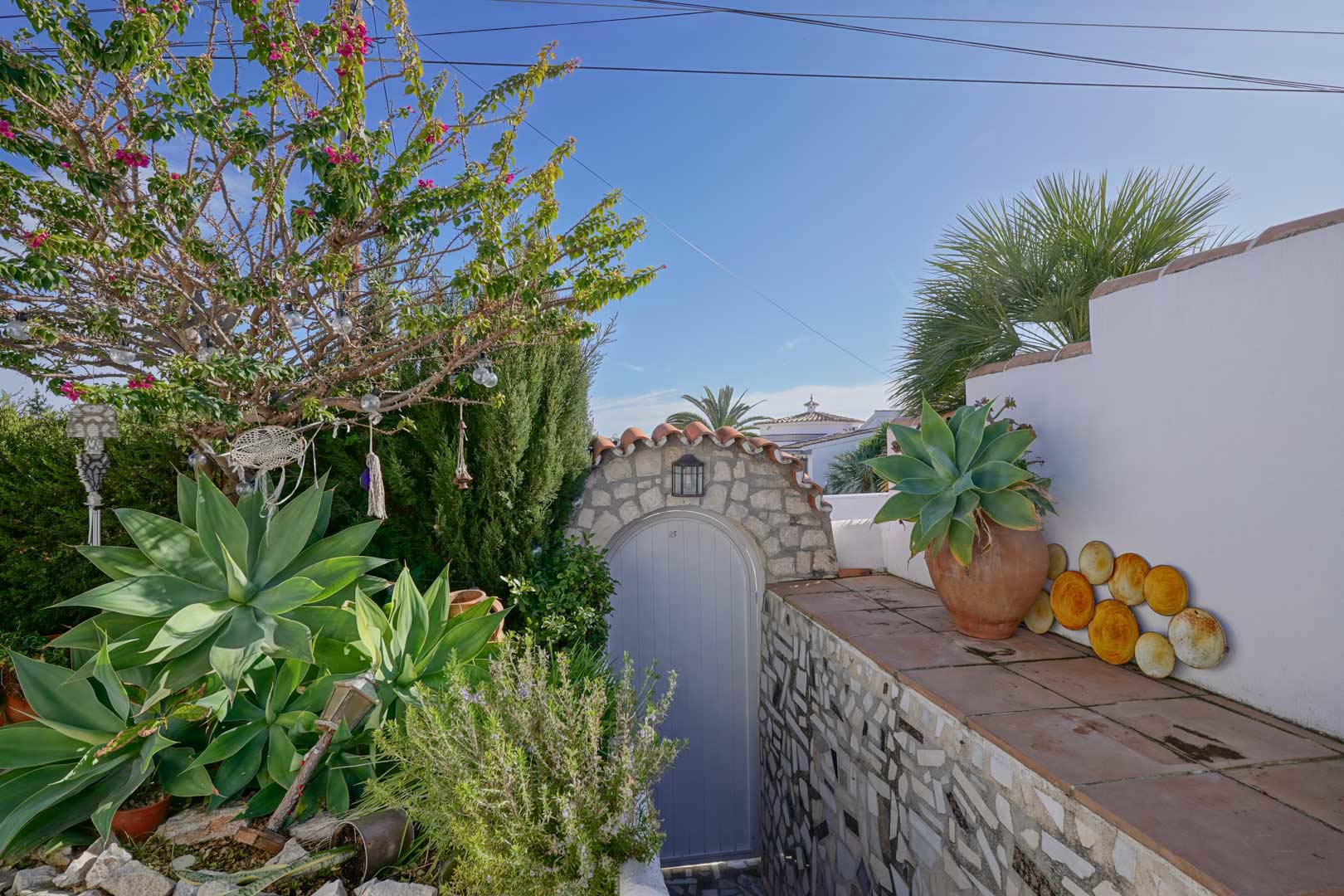 Außenbereich mit Natursteinbogen, hellem Tor und mediterraner Bepflanzung wie Agaven und Bougainvillea vor weiß getünchten Mauern und Terrakottafliesen.