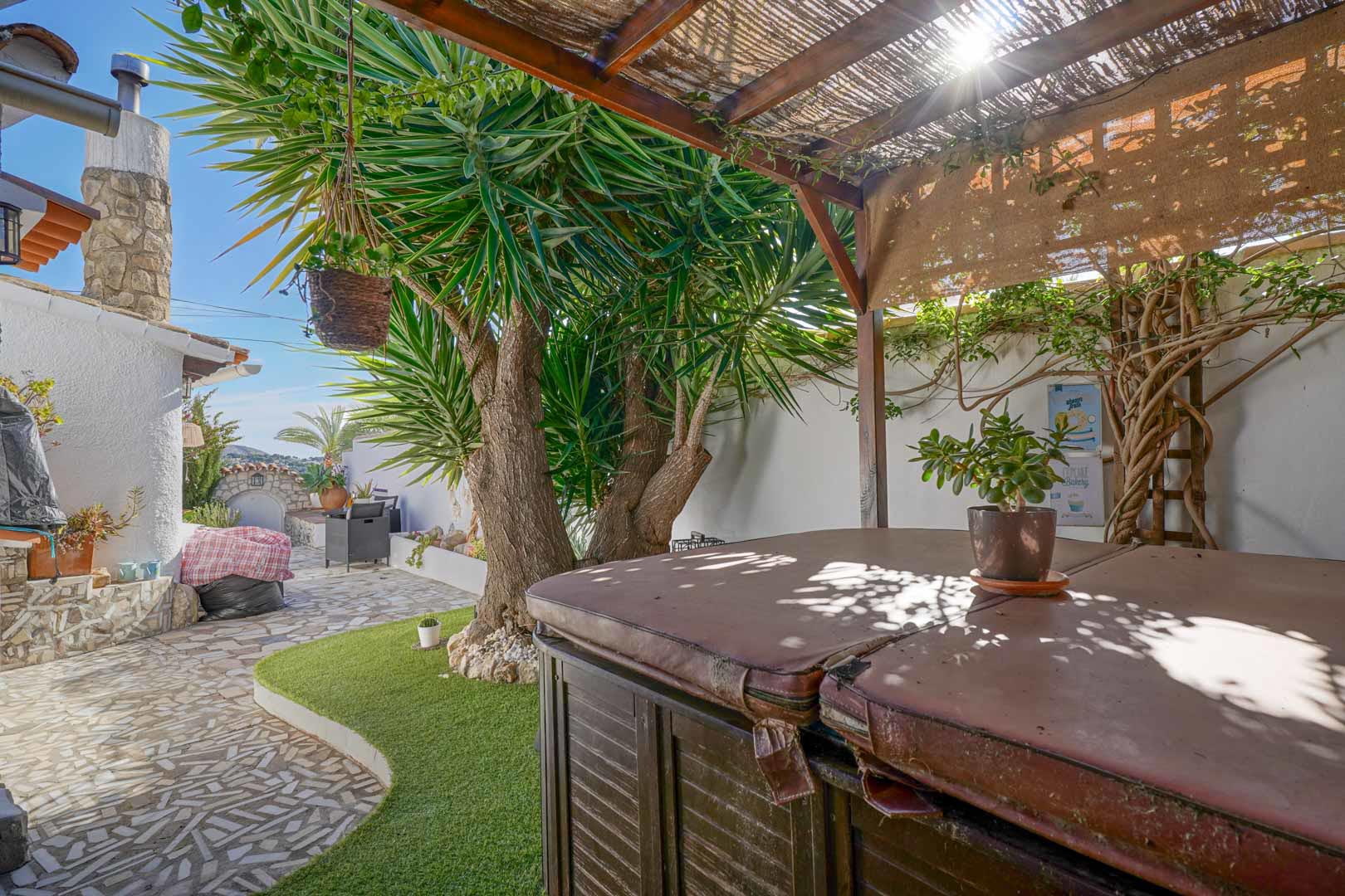 Außenterrasse mit überdachtem Jacuzzi unter einer Holzpergola, eingebettet in einen pflegeleichten Garten mit Steinpflaster und mediterranen Pflanzen.