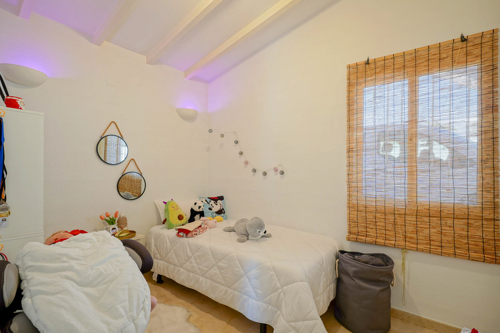 Gemütliches Schlafzimmer mit weiß gestrichener Balkendecke, Einzelbett, dekorativen Spiegeln und einem Naturfaser-Rollo für ein warmes Ambiente.